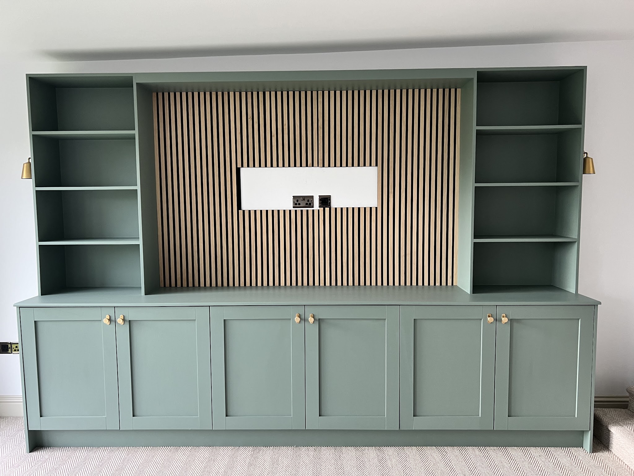 Sage green shelving media unit with a slatted timber panel and brass wall lights